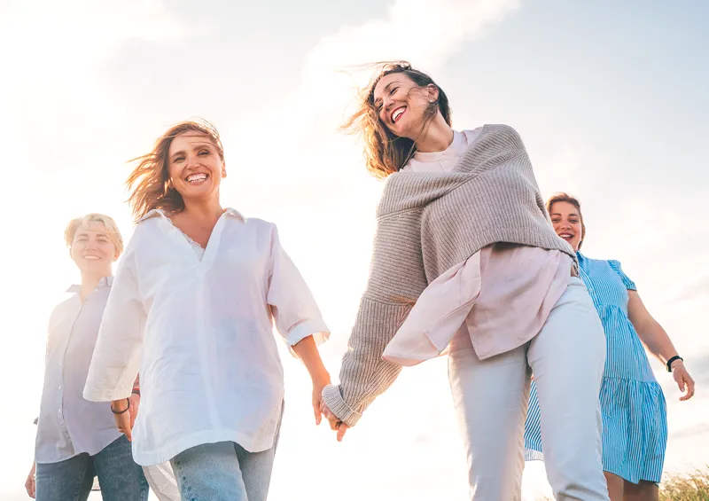 vier Frauen gehen glücklich Hand in Hand, im Hintergrund scheint die Sonne- Symbol für die vier Phasen der Wechseljahre 